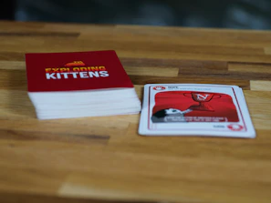 A wooden table features a stack of cards with a red cover labeled 'Exploding Kittens'. Next to it, a single card is laid out showing artwork and text related to the game. The setting is casual and likely related to a tabletop gaming session.