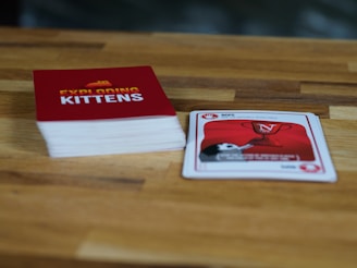 A wooden table features a stack of cards with a red cover labeled 'Exploding Kittens'. Next to it, a single card is laid out showing artwork and text related to the game. The setting is casual and likely related to a tabletop gaming session.