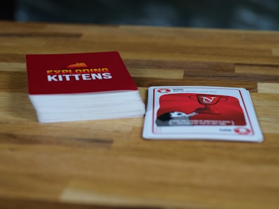 A wooden table features a stack of cards with a red cover labeled 'Exploding Kittens'. Next to it, a single card is laid out showing artwork and text related to the game. The setting is casual and likely related to a tabletop gaming session.