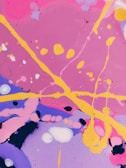Vibrant abstract art featuring swirling patterns and splatters of paint. Bright pink, purple, and blue backgrounds are intersected by bold yellow lines and various circular or amorphous forms. The composition is lively and dynamic, with a playful arrangement of colors and shapes.