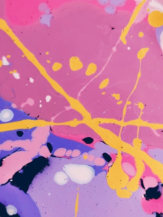 Vibrant abstract art featuring swirling patterns and splatters of paint. Bright pink, purple, and blue backgrounds are intersected by bold yellow lines and various circular or amorphous forms. The composition is lively and dynamic, with a playful arrangement of colors and shapes.