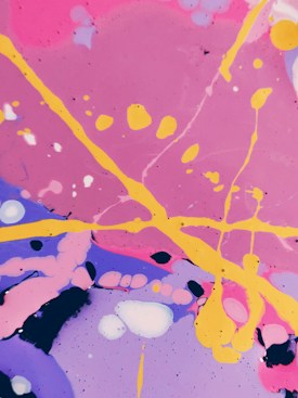 Vibrant abstract art featuring swirling patterns and splatters of paint. Bright pink, purple, and blue backgrounds are intersected by bold yellow lines and various circular or amorphous forms. The composition is lively and dynamic, with a playful arrangement of colors and shapes.