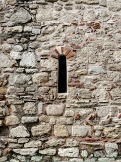 A stone wall with various sizes and shapes of stones tightly fitted together. In the middle, there is a narrow, arched window with a dark interior, framed by reddish-brown bricks. The overall texture is rough and uneven, with hints of mortar visible between the stones.