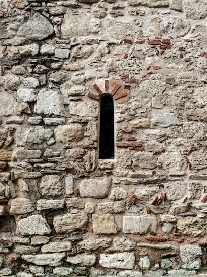 A stone wall with various sizes and shapes of stones tightly fitted together. In the middle, there is a narrow, arched window with a dark interior, framed by reddish-brown bricks. The overall texture is rough and uneven, with hints of mortar visible between the stones.