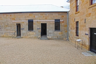 A prison courtyard with stone buildings featuring barred windows and open doorways. The ground is covered with gravel, and there is a small sign on the right side. Metal railings accompany the steps leading to each doorway.