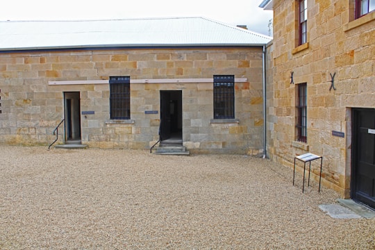 A prison courtyard with stone buildings featuring barred windows and open doorways. The ground is covered with gravel, and there is a small sign on the right side. Metal railings accompany the steps leading to each doorway.