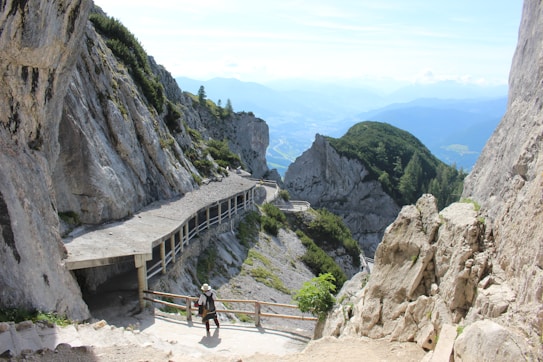 A rugged mountain pathway bordered by steep rock faces and lush greenery extends into a scenic valley. A lone hiker wearing a hat is captured mid-step, drawing attention to the sheer scale of the natural surroundings. The distant mountains and sky create a serene backdrop.