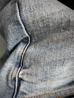 Close-up view of denim fabric showcasing the texture and stitching details. The fabric appears to be part of a garment, possibly jeans, with visible seams and traditional blue coloration.