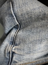 Close-up view of denim fabric showcasing the texture and stitching details. The fabric appears to be part of a garment, possibly jeans, with visible seams and traditional blue coloration.