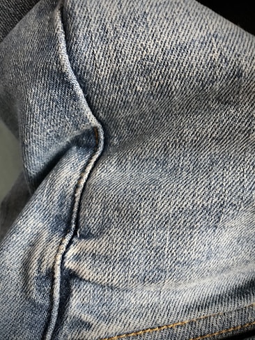 Close-up view of denim fabric showcasing the texture and stitching details. The fabric appears to be part of a garment, possibly jeans, with visible seams and traditional blue coloration.