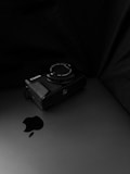 A digital camera rests on top of a closed laptop. The camera is positioned predominantly in the image while the laptop's Apple logo is clearly visible. The background is mostly dark, providing a contrast that highlights the objects.