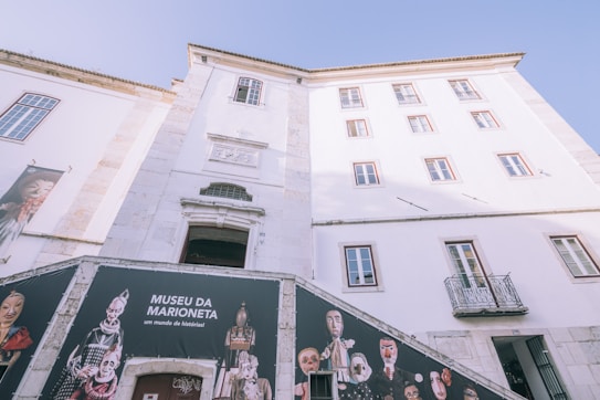 A tall, white building with multiple windows and a classical architectural style. The banner on the lower left features puppets and the text 'Museu da Marioneta' next to them. Some balconies are visible, and the sky is clear and blue, contributing to the bright appearance of the scene.
