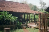 A rustic wooden structure with a tiled roof sits surrounded by greenery. The setting includes a bamboo fence, wooden chairs, and an outdoor lamp. The atmosphere is calm and natural, featuring lush plants and a serene outdoor environment.