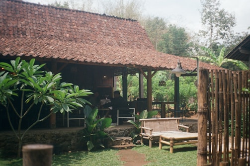 A rustic wooden structure with a tiled roof sits surrounded by greenery. The setting includes a bamboo fence, wooden chairs, and an outdoor lamp. The atmosphere is calm and natural, featuring lush plants and a serene outdoor environment.