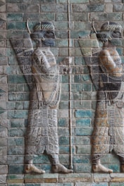 The image features a bas-relief of ancient soldiers adorned with intricate patterns, holding spears and shields. The artwork is set on a backdrop of colored tiles, exhibiting detailed craftsmanship and historical significance.