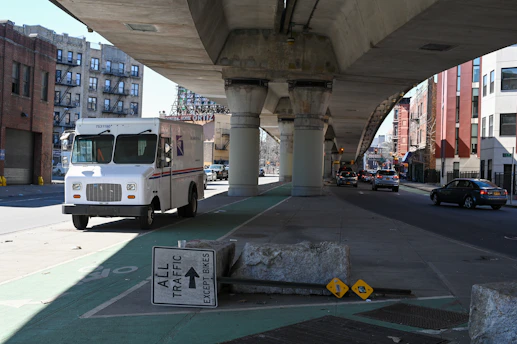A large postal delivery truck is parked on a shaded street under an elevated railway. Nearby, traffic signage reads 'All Traffic Except Bikes', and there is a concrete barrier. The street is lined with a mix of residential and commercial buildings, and there are cars driving on the road in the background.