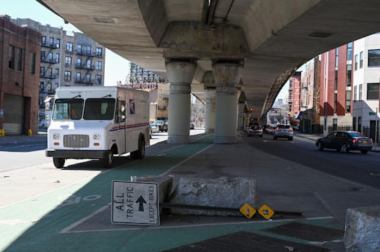 A large postal delivery truck is parked on a shaded street under an elevated railway. Nearby, traffic signage reads 'All Traffic Except Bikes', and there is a concrete barrier. The street is lined with a mix of residential and commercial buildings, and there are cars driving on the road in the background.