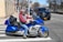 A person wearing a helmet and a red and gray jacket is riding a blue motorcycle at a city intersection. Several cars, including a black SUV, line up on the road in the background. The setting appears urban, with buildings, sidewalks, and trees visible.