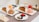 A table set with two rectangular plates each holding a slice of layered cake paired with different ice cream scoops. One plate has chocolate ice cream while the other has strawberry ice cream. In the foreground, there are small pastries with colorful toppings on a round plate. A patterned table runner adds a decorative touch. An orange rose, a red rose, and a small candle in a glass holder are also placed on the table.
