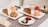 A table set with two rectangular plates each holding a slice of layered cake paired with different ice cream scoops. One plate has chocolate ice cream while the other has strawberry ice cream. In the foreground, there are small pastries with colorful toppings on a round plate. A patterned table runner adds a decorative touch. An orange rose, a red rose, and a small candle in a glass holder are also placed on the table.