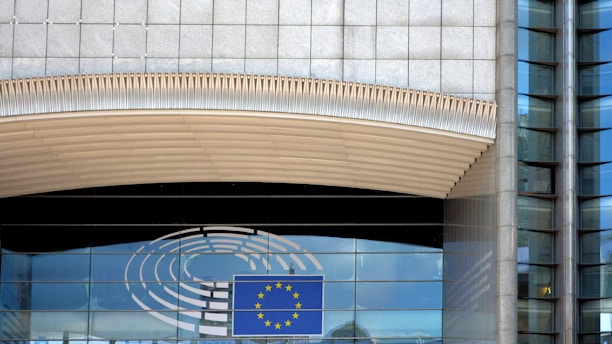 A modern building facade with a large window reflecting the sky. An emblem with a circular design and a blue rectangle featuring yellow stars, reminiscent of the European Union flag, is visible. The architecture includes glass panels and vertical lines with metallic tones.