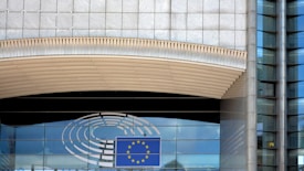 A modern building facade with a large window reflecting the sky. An emblem with a circular design and a blue rectangle featuring yellow stars, reminiscent of the European Union flag, is visible. The architecture includes glass panels and vertical lines with metallic tones.