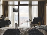 A cozy bedroom with a bed facing large windows that reveal a cityscape. The room features soft lighting, plush curtains, and neutral tones. There is a small table with a cap and some items on it, a comfortable chair, and a tall floor lamp. A person’s feet in socks are visible, suggesting they are relaxing on the bed.