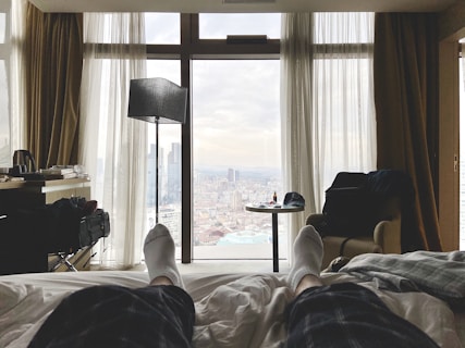 A cozy bedroom with a bed facing large windows that reveal a cityscape. The room features soft lighting, plush curtains, and neutral tones. There is a small table with a cap and some items on it, a comfortable chair, and a tall floor lamp. A person&rsquo;s feet in socks are visible, suggesting they are relaxing on the bed.