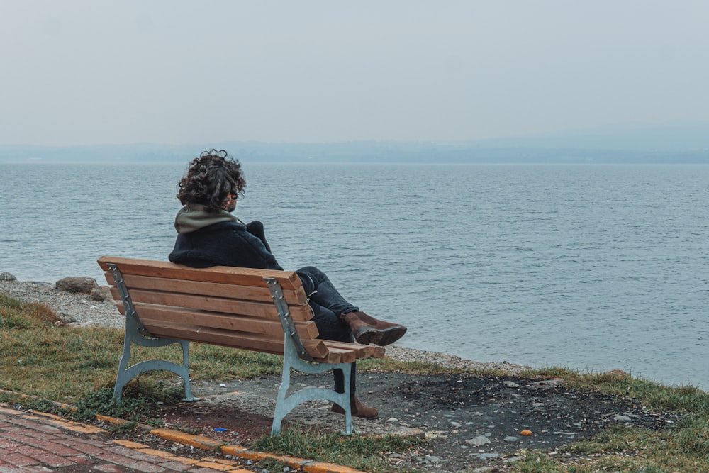 woman sitting on brown wooden bench near body of water during daytime