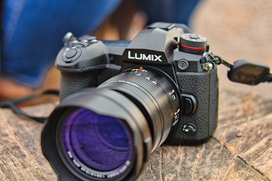 A Lumix camera rests on a textured wooden surface. The camera has a lens with a UV filter, and there is a prominent red accent near the shutter button. The camera body features a textured grip and various buttons and dials for control.