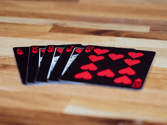 A set of playing cards is spread out on a wooden surface, featuring a royal flush in hearts. The cards are black with red heart symbols and include the Ace, King, Queen, Jack, and ten of hearts.