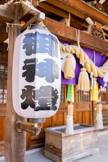 A traditional Japanese paper lantern with kanji characters is hanging in front of a wooden structure. Behind the lantern, a purple cloth decorated with large, twisted straw ropes with tassels is visible. A wooden offering box is positioned below the lantern.