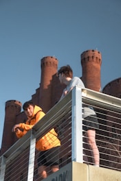 Two young men are standing on an elevated platform with a metallic railing, against the backdrop of a large brick building featuring multiple round towers. One of the individuals is wearing a bright orange jacket, while the other is dressed in a gray shirt and black shorts. They appear to be engaged in conversation.