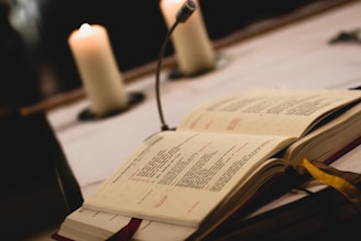 An open book with printed text lies on a table, next to a vertical microphone. Two lit candles are softly illuminated in the background, creating a warm ambiance.