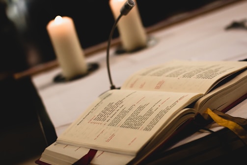 An open book with printed text lies on a table, next to a vertical microphone. Two lit candles are softly illuminated in the background, creating a warm ambiance.