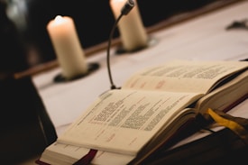 An open book with printed text lies on a table, next to a vertical microphone. Two lit candles are softly illuminated in the background, creating a warm ambiance.
