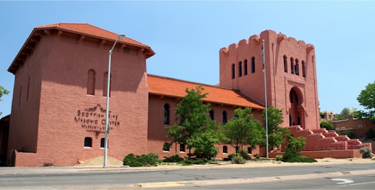 A large building with a reddish-brown facade and architectural features resembling a fortress or castle. It has a prominent tower with a series of arched windows and an entrance accentuated by a grand archway. The structure is surrounded by greenery, including a few small trees and bushes. An American flag is visible on a flagpole, and the building's sign identifies it as a Masonic center.