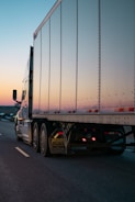 A large semi-trailer truck with a shiny metallic trailer is driving along a highway. The road appears to be open and mostly empty, suggesting a long-distance journey. The sky is clear with a gradient from warm orange to cool blue, indicating either sunrise or sunset.