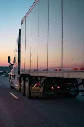 A large semi-trailer truck with a shiny metallic trailer is driving along a highway. The road appears to be open and mostly empty, suggesting a long-distance journey. The sky is clear with a gradient from warm orange to cool blue, indicating either sunrise or sunset.