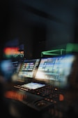 A dimly lit control room featuring multiple computer screens filled with colorful graphics and data, creating an atmosphere of high-tech operations. There is a keyboard in the foreground, and a digital clock display is visible showing a time countdown.