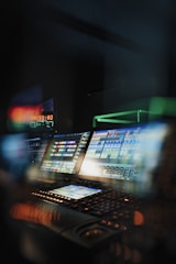 A dimly lit control room featuring multiple computer screens filled with colorful graphics and data, creating an atmosphere of high-tech operations. There is a keyboard in the foreground, and a digital clock display is visible showing a time countdown.
