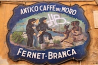 A vintage-style signboard with the text 'Antico Caffè del Moro Fernet-Branca' against a weathered wall. The sign features a painted illustration of several people, some holding bottles, dressed in historical clothing. Two pairs of men and women can be seen interacting.