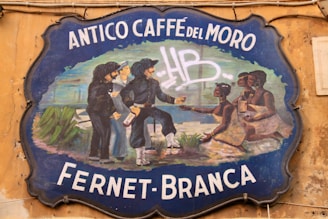 A vintage-style signboard with the text 'Antico Caffè del Moro Fernet-Branca' against a weathered wall. The sign features a painted illustration of several people, some holding bottles, dressed in historical clothing. Two pairs of men and women can be seen interacting.