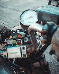 A close-up view of a motorcycle's dashboard featuring a tachometer, several indicators, and a metal handlebar. The tachometer displays ranges from 0 to 11, marked at intervals, while the indicator lights below are covered in transparent panels of various colors. The metal parts exhibit signs of wear, adding to the rugged aesthetic.