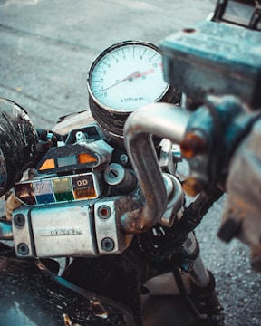 A close-up view of a motorcycle's dashboard featuring a tachometer, several indicators, and a metal handlebar. The tachometer displays ranges from 0 to 11, marked at intervals, while the indicator lights below are covered in transparent panels of various colors. The metal parts exhibit signs of wear, adding to the rugged aesthetic.