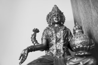 A detailed bronze statue of a deity sitting cross-legged. The statue features intricate designs on the clothing and jewelry. The figure holds a flower in one hand and an ornate vessel in the other. The background is plain, emphasizing the statue's features.