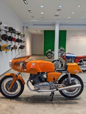 A classic orange motorcycle is prominently displayed indoors, surrounded by various vintage helmets mounted on the wall. In the background, there are more motorcycles, and a sign on the wall reads 'Stuart Parr Collection'. The setting appears to be a showroom or exhibition space, with polished concrete floors and track lighting on the ceiling.