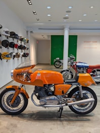A classic orange motorcycle is prominently displayed indoors, surrounded by various vintage helmets mounted on the wall. In the background, there are more motorcycles, and a sign on the wall reads 'Stuart Parr Collection'. The setting appears to be a showroom or exhibition space, with polished concrete floors and track lighting on the ceiling.