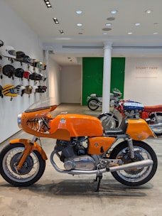 A classic orange motorcycle is prominently displayed indoors, surrounded by various vintage helmets mounted on the wall. In the background, there are more motorcycles, and a sign on the wall reads 'Stuart Parr Collection'. The setting appears to be a showroom or exhibition space, with polished concrete floors and track lighting on the ceiling.