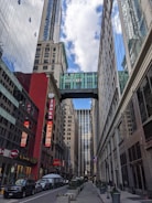 A city street with tall buildings on both sides. A skybridge connects two buildings at an upper level near the top of the frame. The buildings are a mix of modern glass and older styles; one building has a red façade with signs advertising a 99 cent store and a restaurant named Jacks. Several vehicles including cars and vans are parked along the street. There are a few pedestrians on the sidewalks.